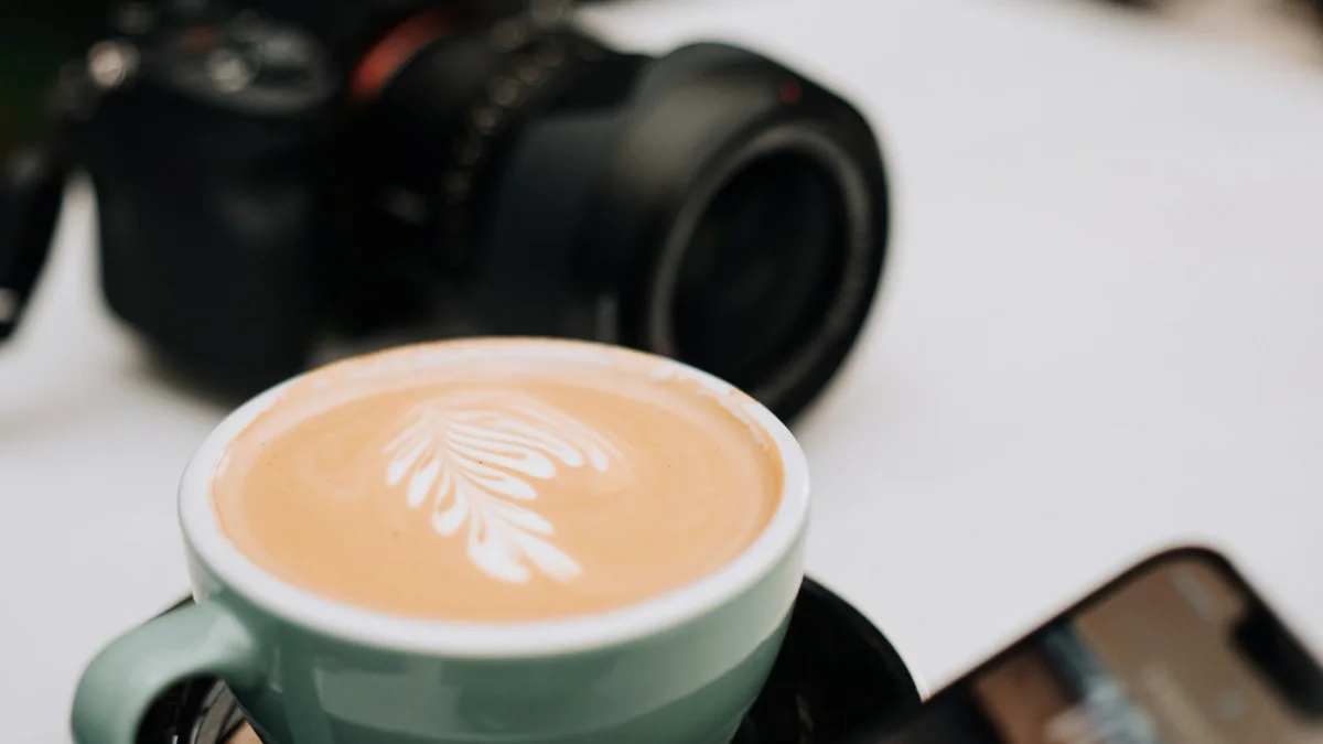 Beyaz masada latte, arkada bir fotoğraf makinesi, sağ altta bir telefon
