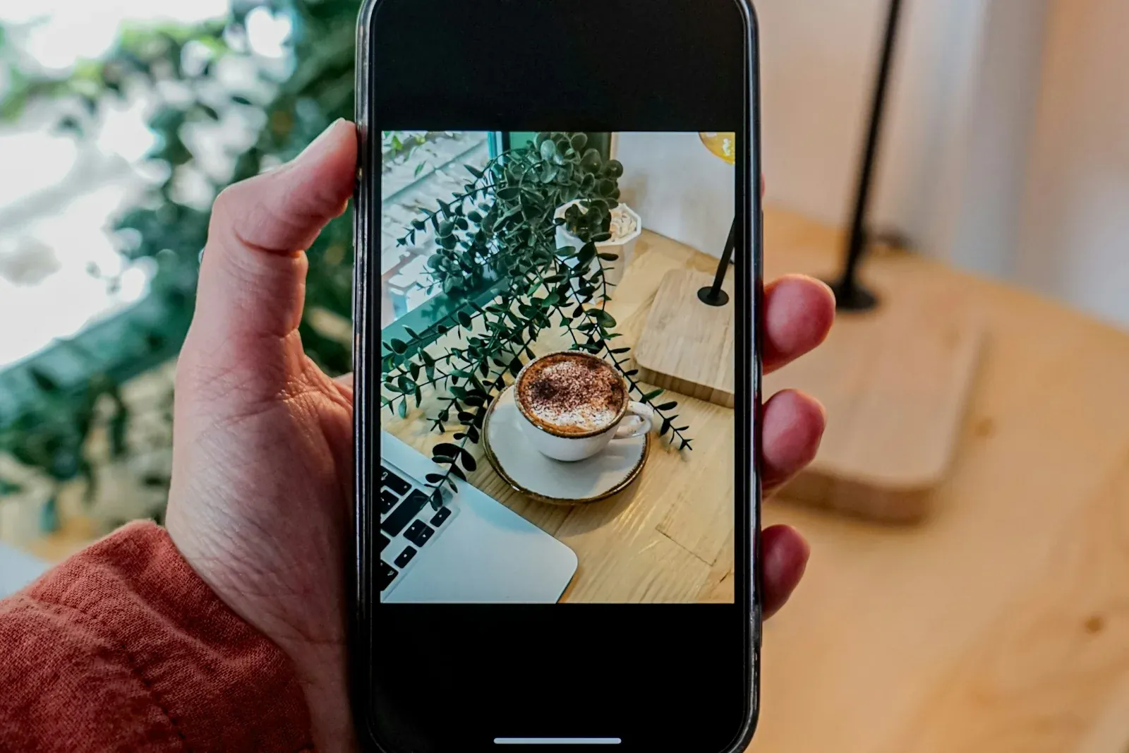 Ahşap masada kahve fincanını dikey tutulan telefonla fotoğraflayan bir el; ekrandaki fotoğrafın üstünde ve altında siyah boşluk var