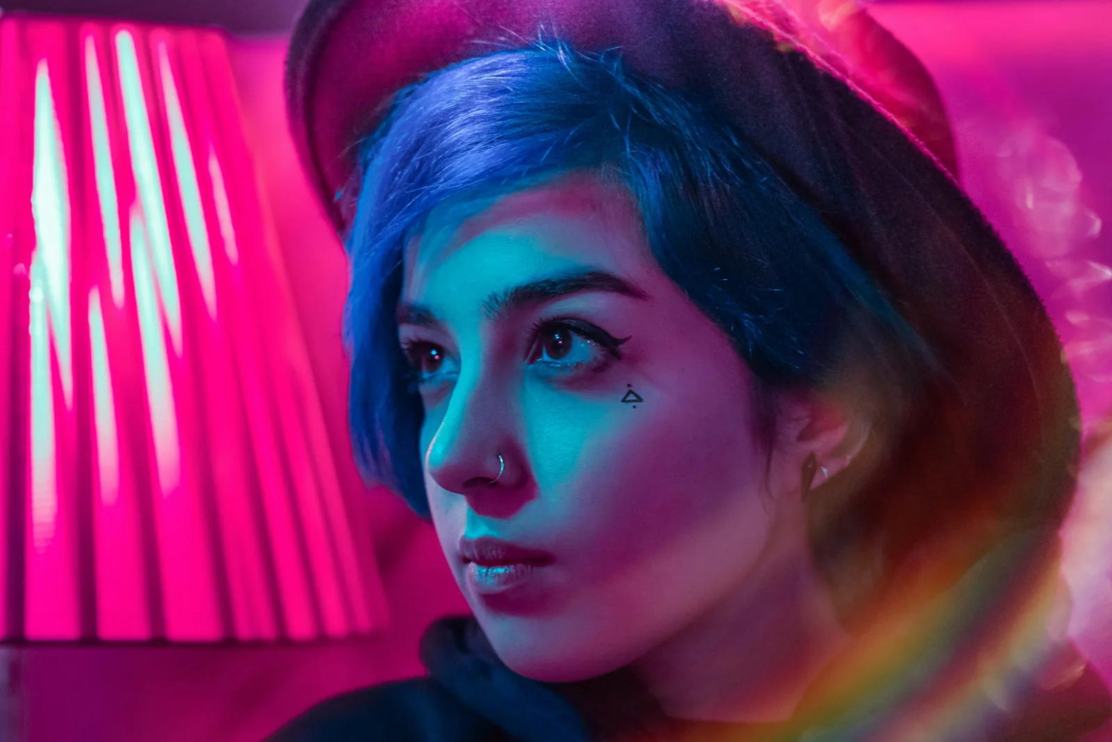 Pembe ve turkuaz ışıkta, şapkalı ve mavi saçlı bir kadının yana bakan yakın çekimi