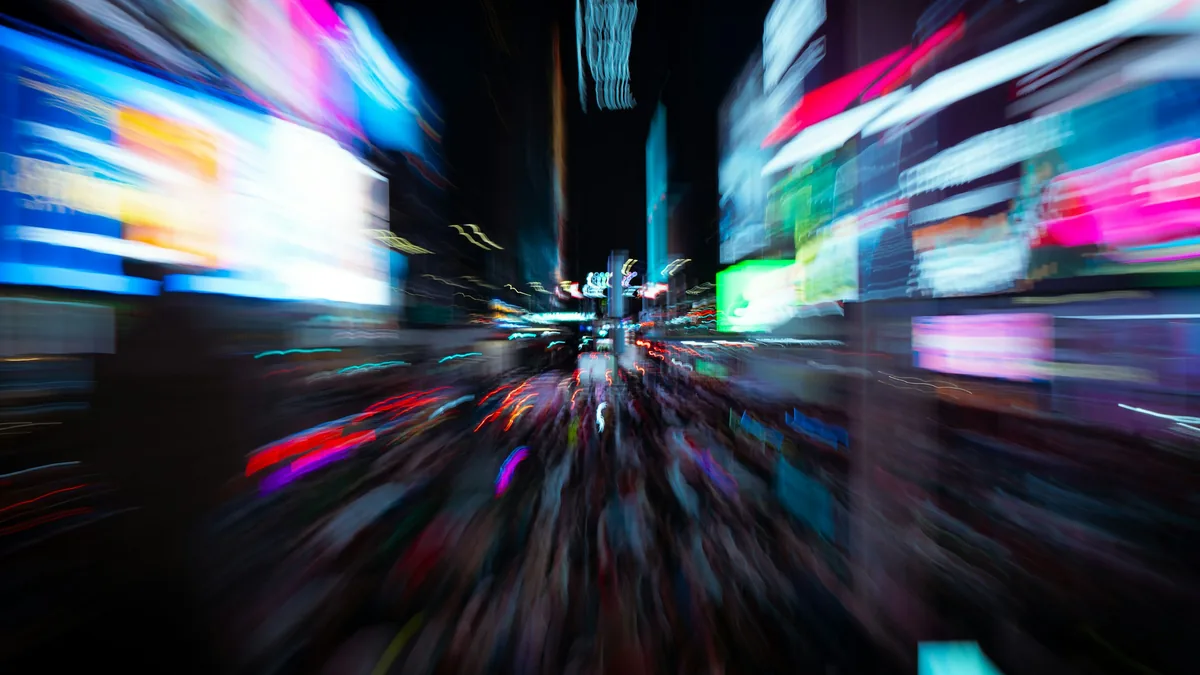 Gece çekilmiş, ışıkları hareketle bulanıklaşmış reklam ekranlarıyla dolu bir şehir caddesi